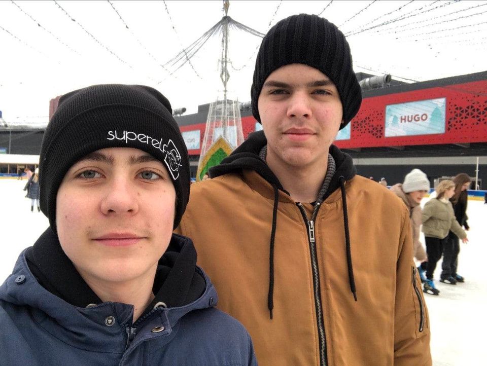 To gutte-ungdommer står ute på en plass med snø og har på seg svarte luer og ytterjakker.