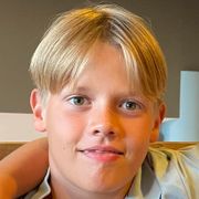 En ung gutt med lyst hår, midtskill-delt pannelugg smiler inn i kameraet.