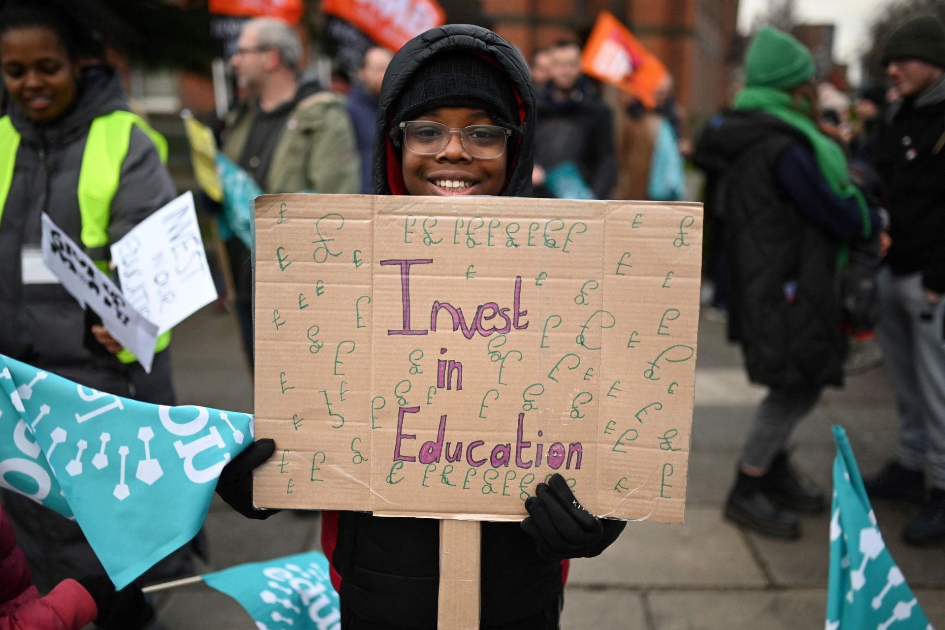 En ung gutt med briller, lue og varm jakke står sammen med mange voksne og holder et pappskilt som det står "Invester i utdanning" på. 