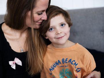En gutt med oransje genser sitter og smiler i en sofa og moren hans sitter ved siden av holder rundt ham og har en rosa sløyfe på brystet. 
