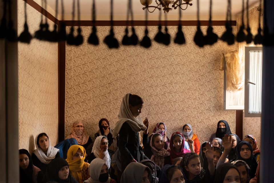 Bildet viser et slags klasserom med tapetserte vegger, der det henger noen objekter i taket, og mange jenter i hijab sitter på gulvet.