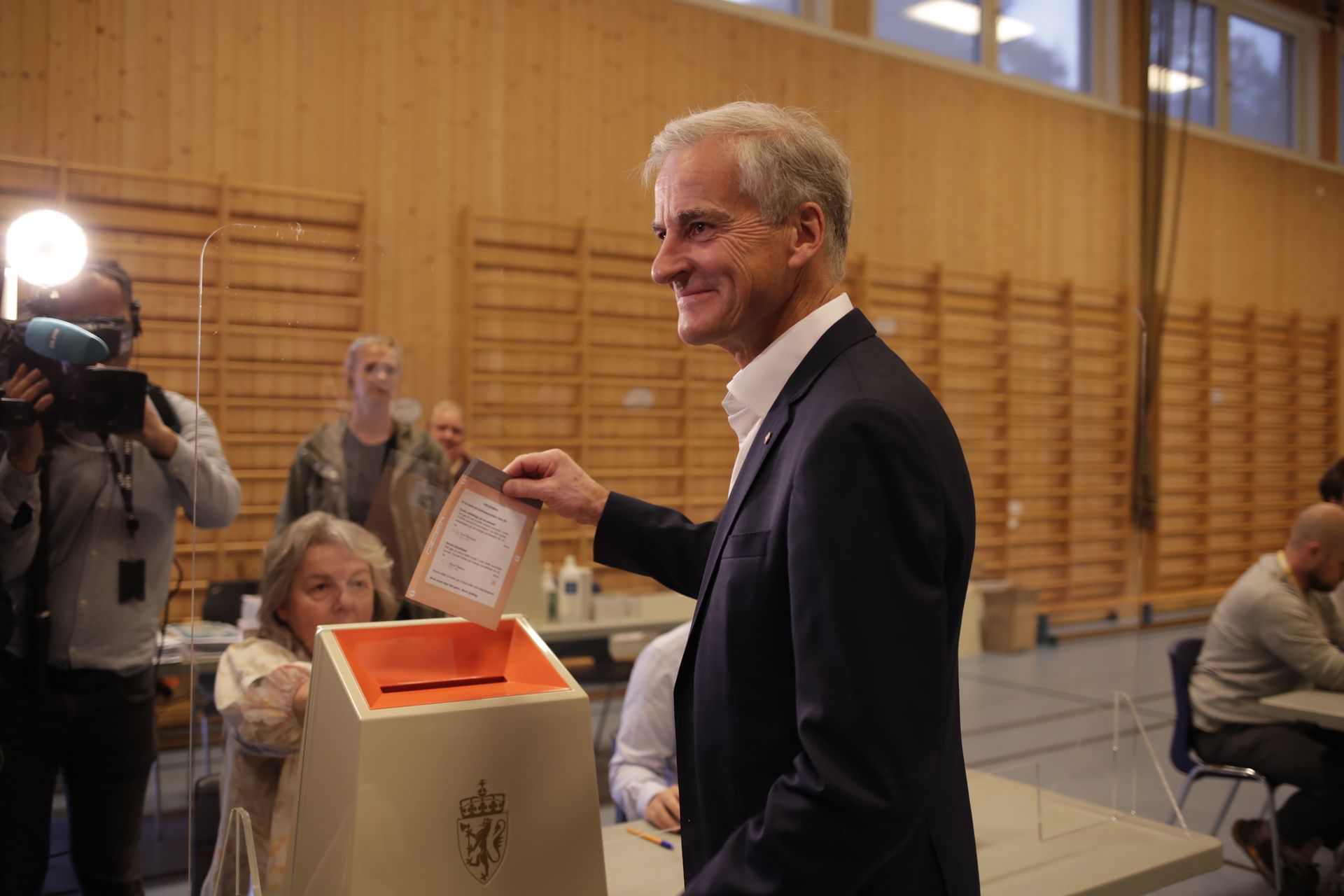 Jonas Gahr Støre smiler mens han stikker en stemmeseddel ned i valgurnen i en gymsal.