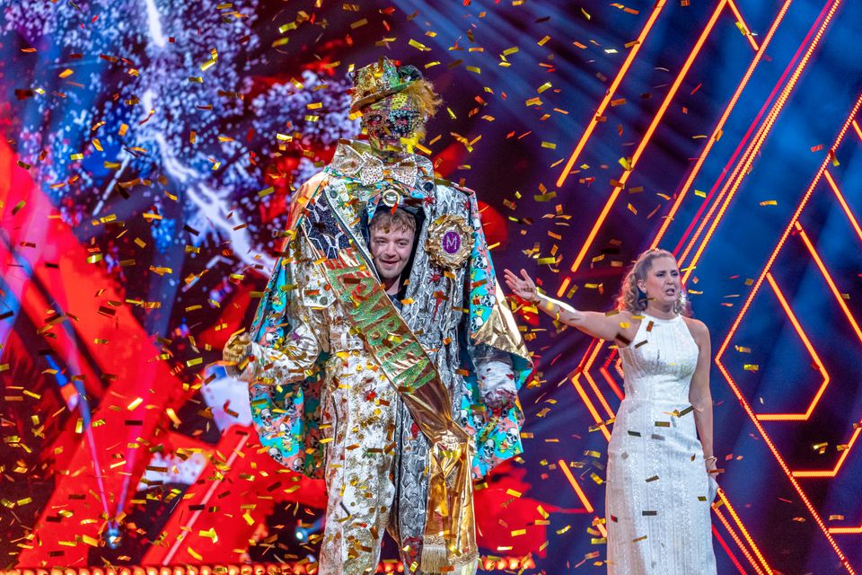 Ansiktet til en gutt kikker ut på midten av et stort zombie-kostyme på en scene ved siden av en kvinne i hvit kjole og det er masse konfetti på scenen. 