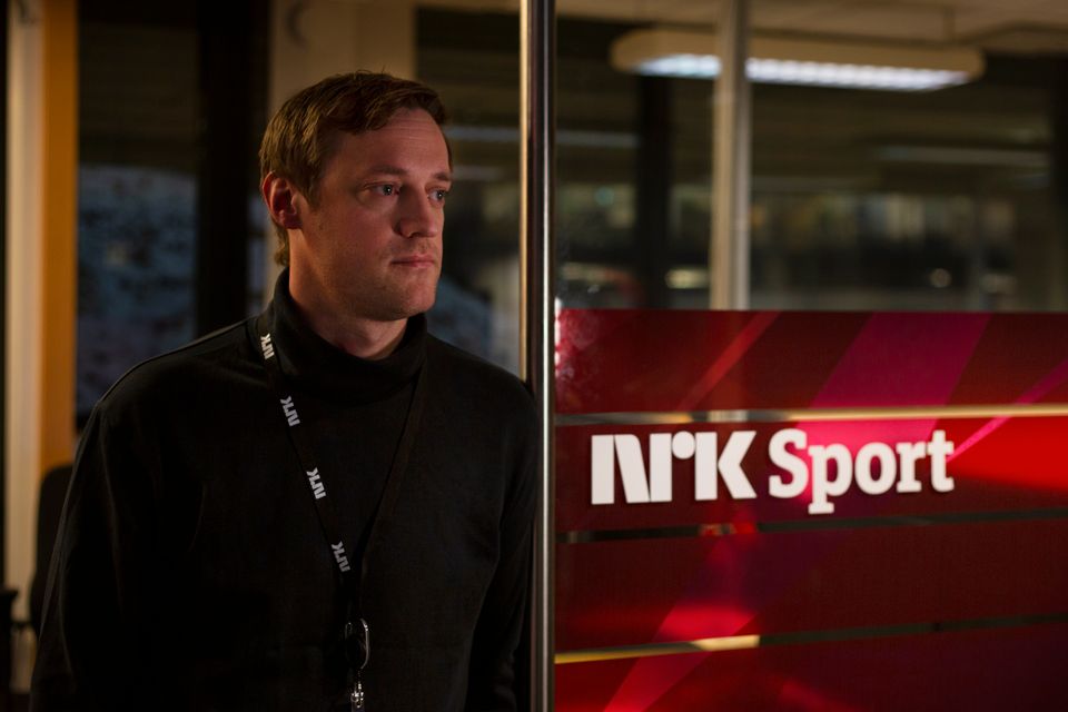 En mann i sort genser lener seg mot en rød vegg hvor det står NRK SPORT med hvite bokstaver.