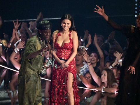 Selena Gomez, en kvinne i 30-årene, står på en scene iført en rød, perlebrodert kjole, sammen med en mann i en grønn type dress og et sølvtrofé i hånden.