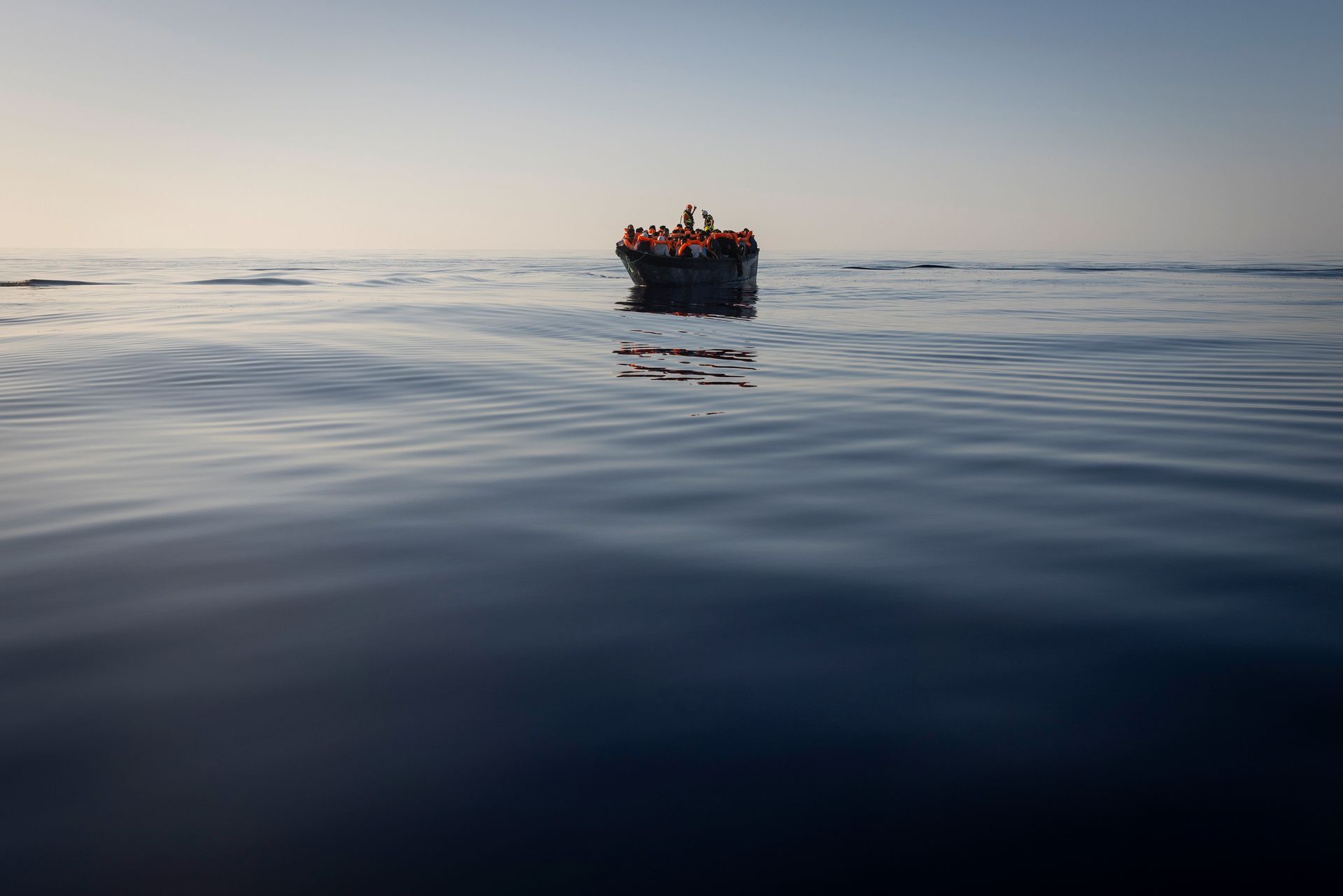 Mange mennesker med oransje redningsvester sitter tett i tett på en liten båt midt ute på havet. 