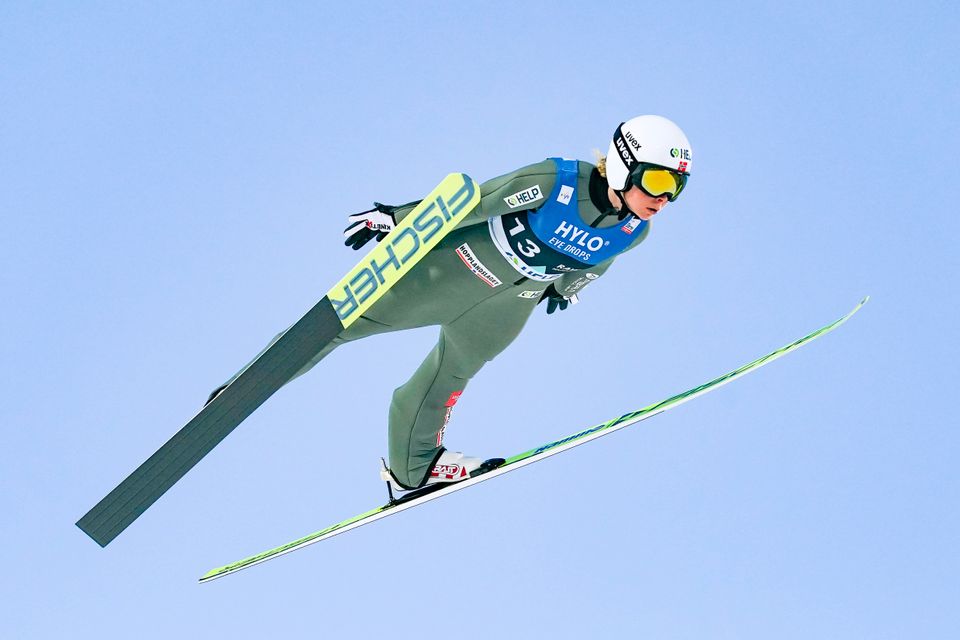 En kvinne svever i luften med lange ski på bena i en grønn trang drakt og hvit hjelm