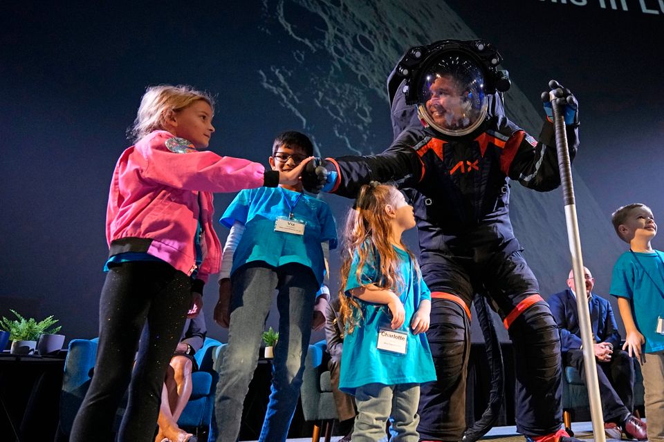 En mann i en sort kjeledress med en gjennomsiktig kule på hodet står på en scene sammen med flere barn og tar den ene jenta i hånda. 