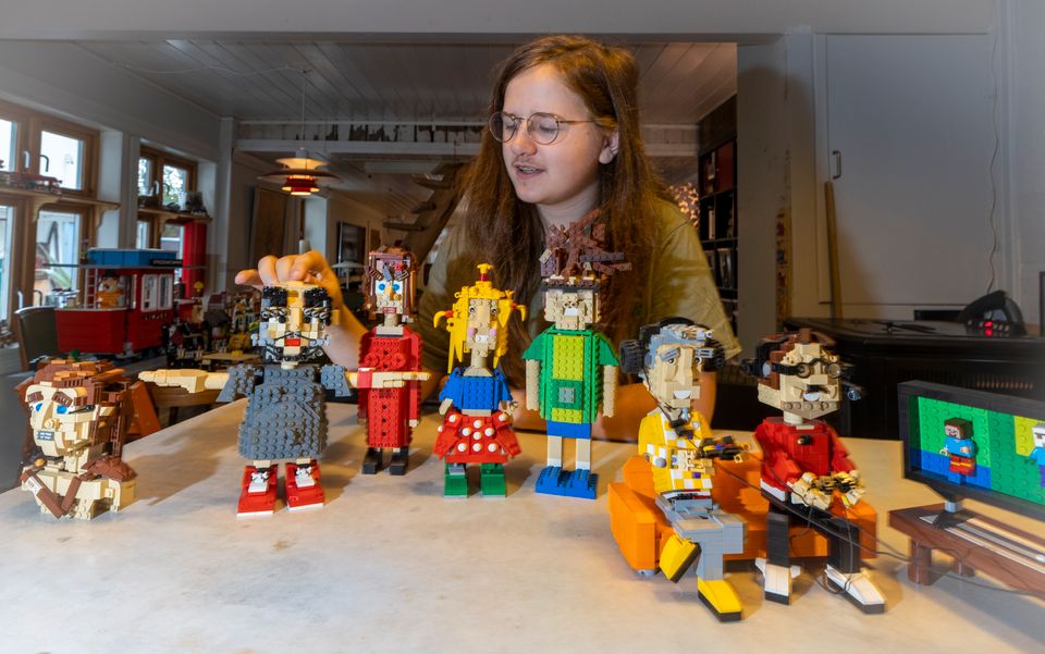 En gutt med briller og langt hår viser frem seks figurer bygget i lego, figurene er av en mann, en gutt, en jente og en dame pluss to gutter som sitter i en sofa og gamer. 