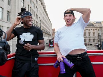 KSI og Logan Paul, to voksne menn, står oppå et rødt lasteplan og kjører rundt i London, mens de holder lilla flasker og blir filmet.