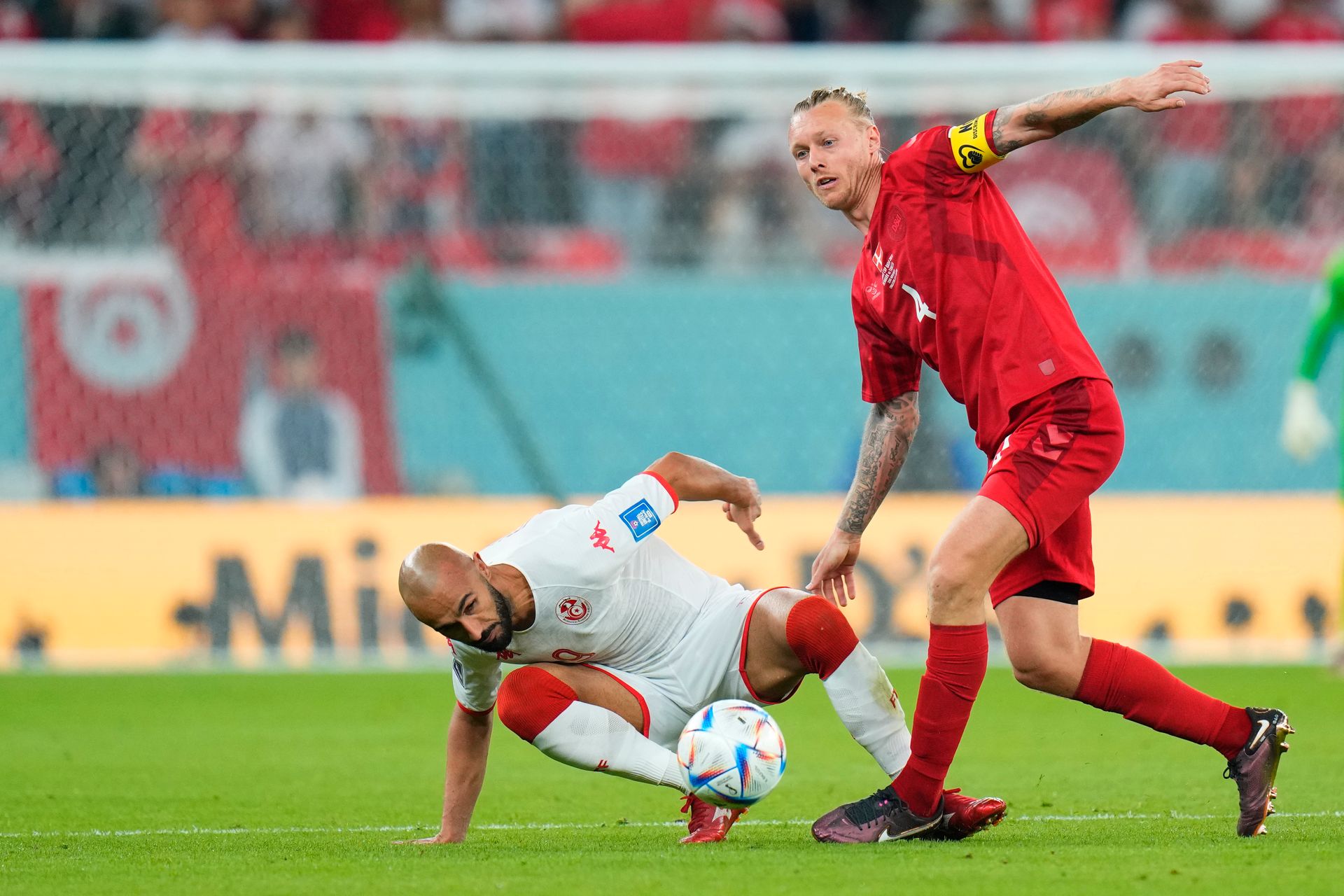 To fotballspillere kjemper om en ball, den ene i rødt står og den andre i hvit drakt ligger på bakken ved siden av.