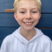 En gutt med lyst hår og hvit hettegenser smiler foran en blå vegg.