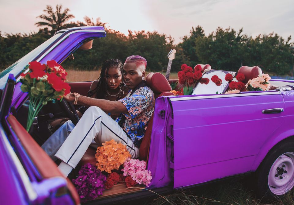 En mann med lilla hår og en dame med mørkt hår sitter i en lilla bil med åpent tak sammen med masse blomster.