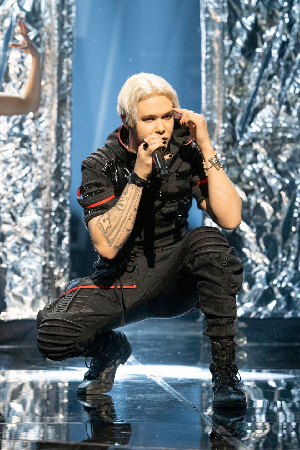 En ung mann med helt lyst hår som er delt i midskill står på en scene og synger inn i en mikrofon og han har satt seg ned på huk. 