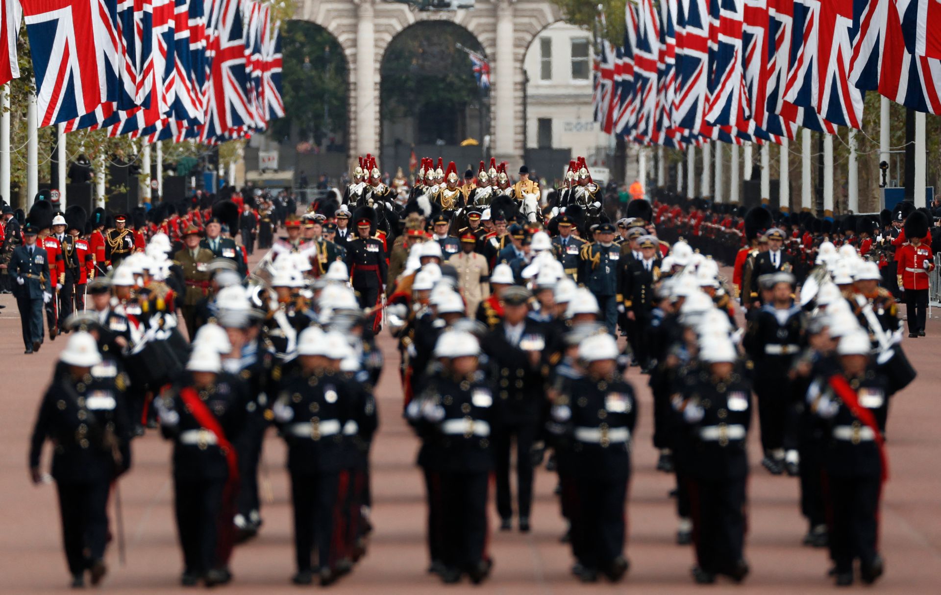 Mange uniformerte menn med hatter på og ryttere bak marsjerer i en bred gate med mange britiske flagg i rødt, hvitt og blått hengende på begge sier av veien. 