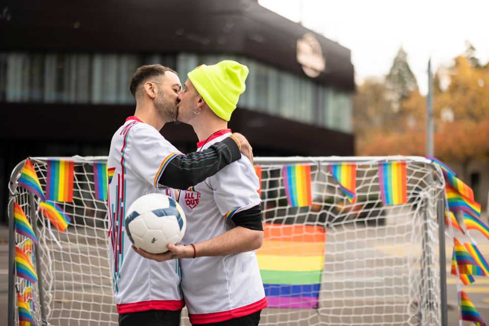 En mann med kort, brunt hår kysser en mann med signalgul lue foran et fotballmål med fargerike flagg og en fotball i hånden.