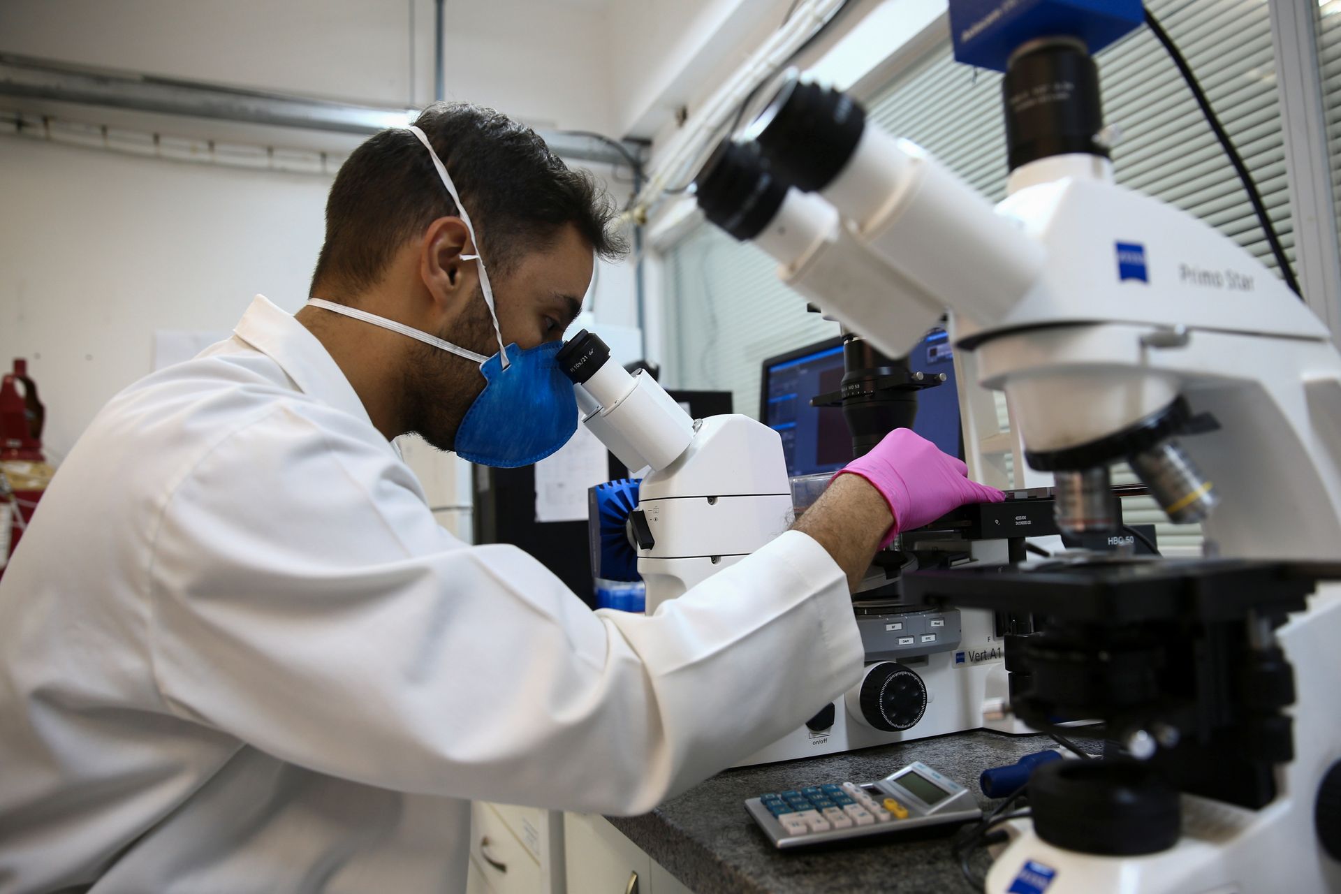 En forsker med hvit frakk og ansiktsmaske studerer en prøve under et mikroskop i et laboratorium. 