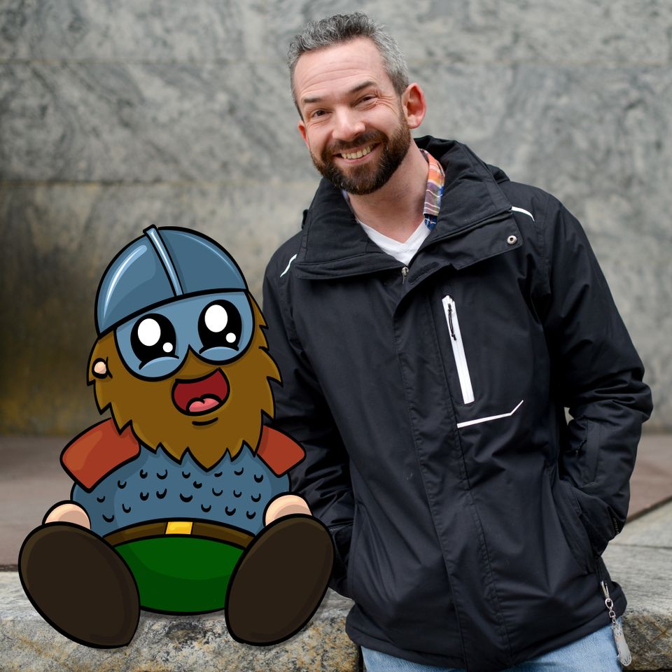 En mann med mørkt skjegg og sort jakke står ved siden av en liten tegneseriefigur som ser ut som en viking med tykt skjegg og vikinghjelm