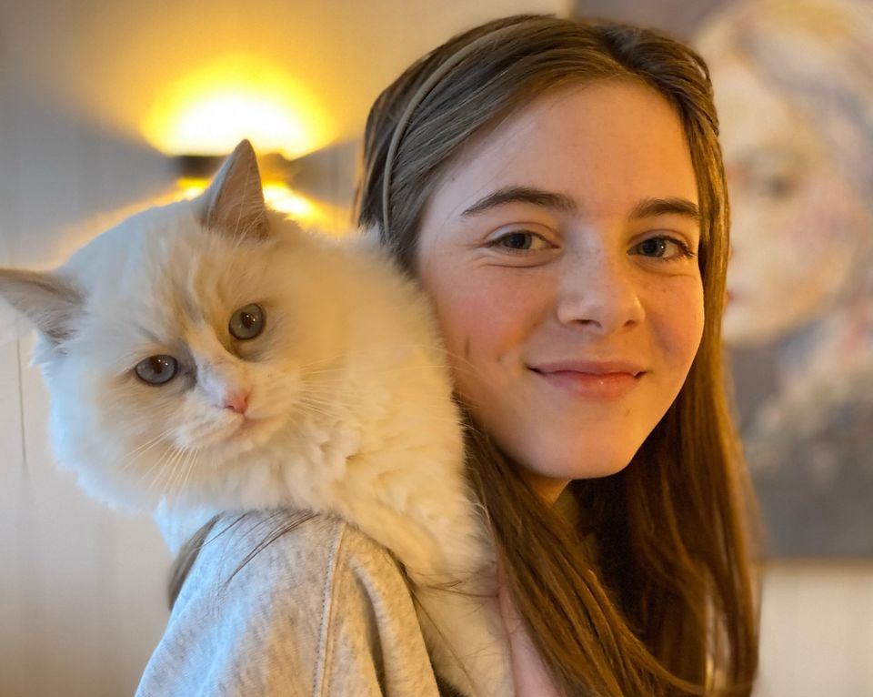 En jente med langt, brunt hår og grå genser smiler og på skulderen hennes er det en hvit katt.
