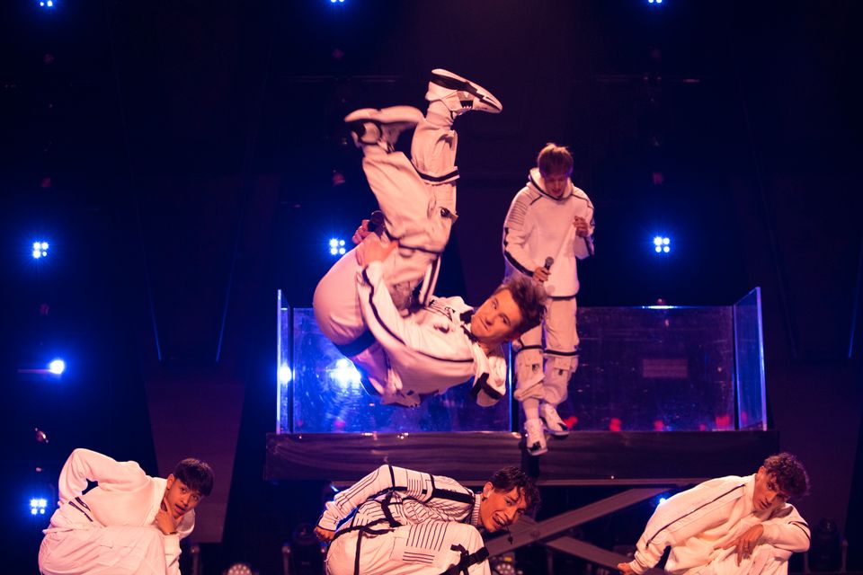 Seks unge menn i hvite klær danser på en scene med blåt lys og den ene tar en salto over de andre. 