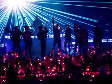 Åtte sangere og en trommeslager står på en scene, mens blått strobelys flommer utover publikummet, som har røde neonlys-ringer rundt håndleddene.