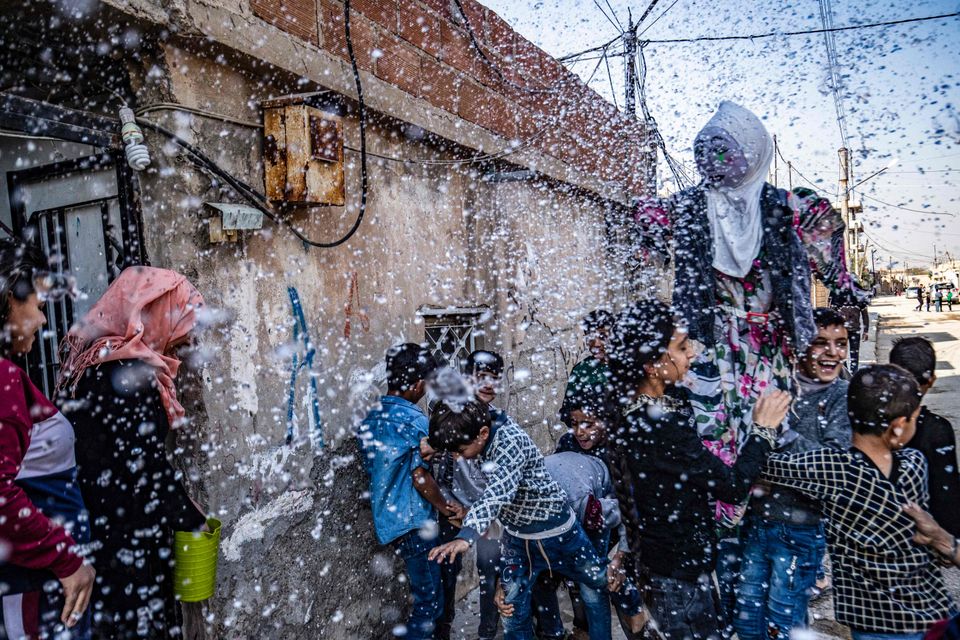 Skum, bobler og vanndråper fyker rundt i luften mens en gruppe barn løfter en stor kvinnedukke med hvitt sjal. 