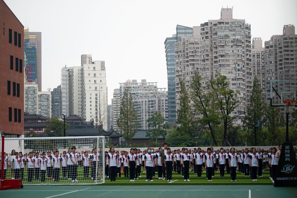 Flere skoleklasser med kinesiske elever i skoleuniformer står oppstilt på rekker på en fotballbane. 