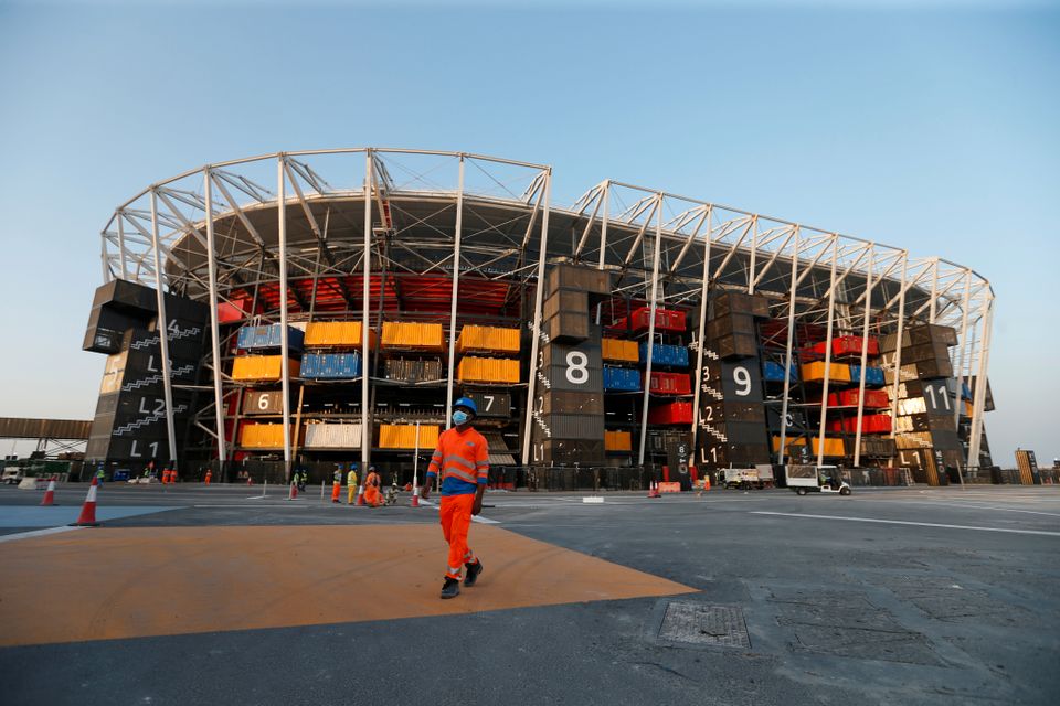 En arbeider i oransje klær går på utsiden av en stor buet fotballstadion som nesten er ferdig bygget.