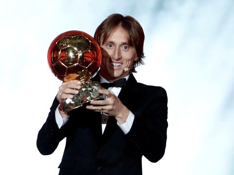 En mann med svart dress, Luka Modric, holder opp et trofé av en fotball i gull.