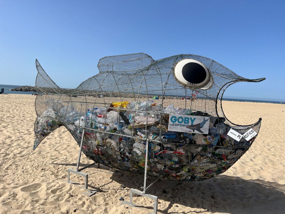 En stor søppelkasse av metall-netting er formet som en gapende fisk, står på en strand og er fylt med plastflasker.