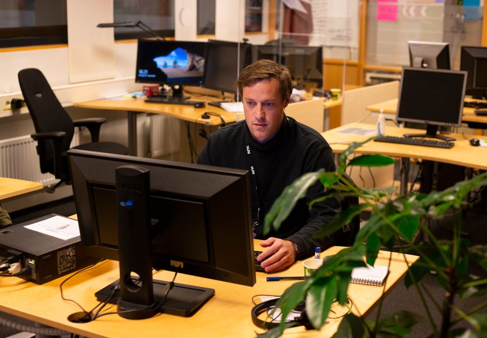 En mann i sort genser sitter ved en pult og jobber på en PC-skjerm i et rom med flere pulter og skjermer. 
