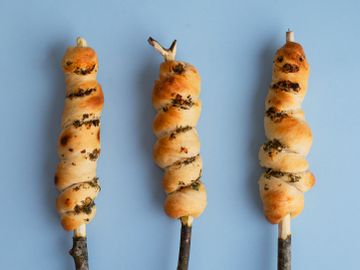 Et bilde tatt ovenfra viser tre gyllent stekte pinnebrød på hver sin spikkede pinne som ligger på en lyseblå bakgrunn.
