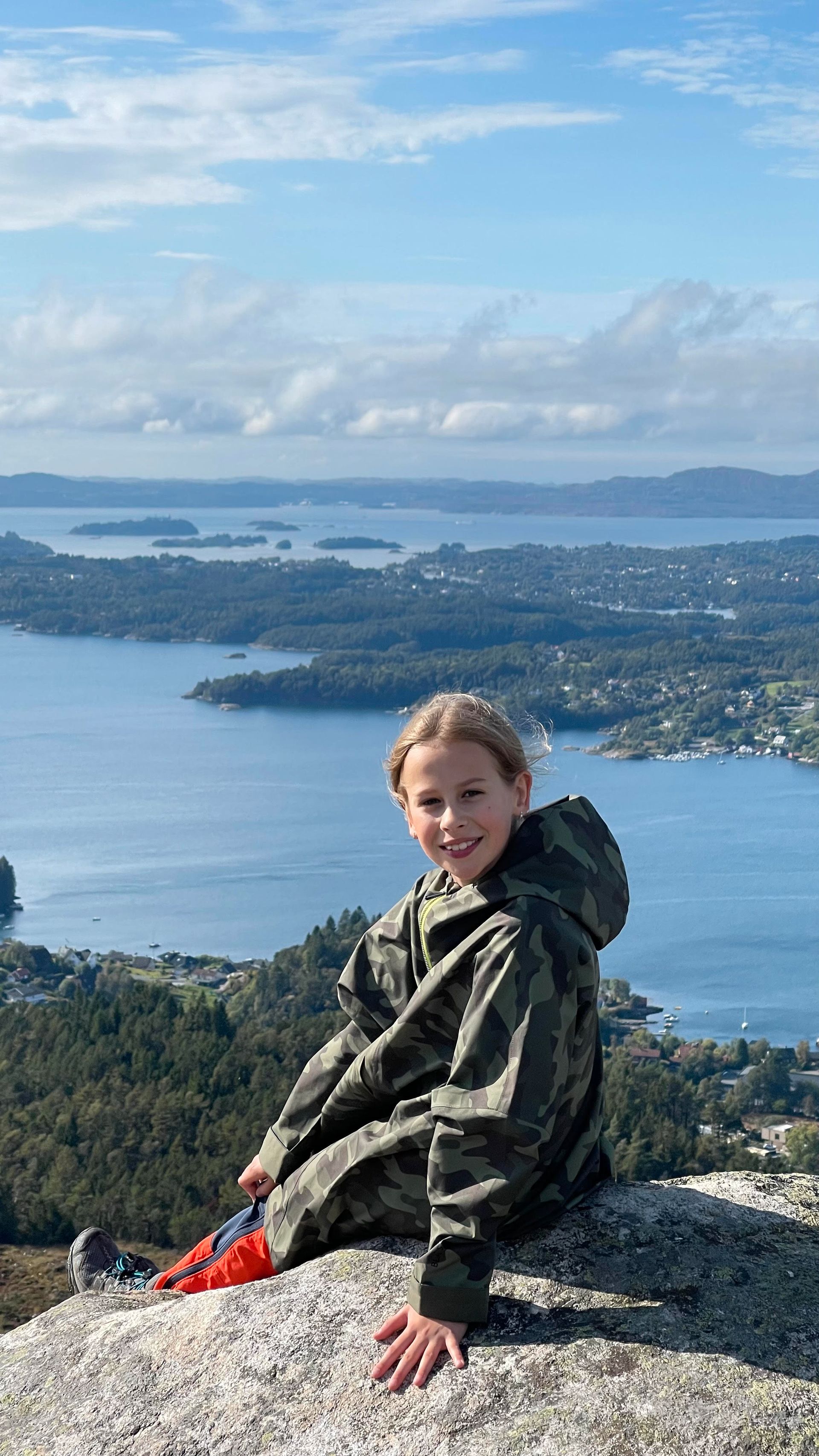 Smilende jente med militærgrønn allværsjakke sitter på en fjelltopp, med en blå fjord i bakgrunnen.