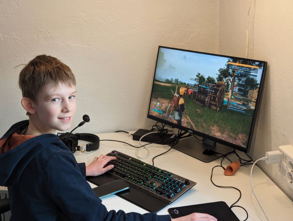 En gutt med mørk hettegenser smiler mot kamera og han sitter foran en pc med et spill på skjermen.