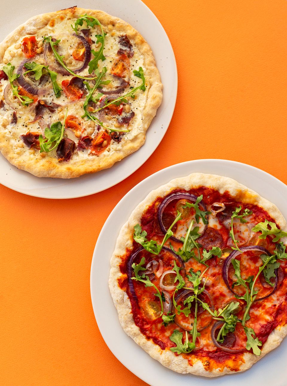 To hvite fat med hver sin pizza oppå, én med hvit saus og én med rød saus, ligger på en knalloransje bakgrunn.