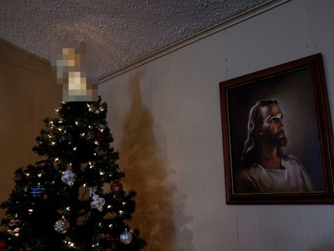 Et juletre med pynt står i et hjørne av et rom, ved siden av et maleri av Jesus, og toppen av juletreet er sladdet.
