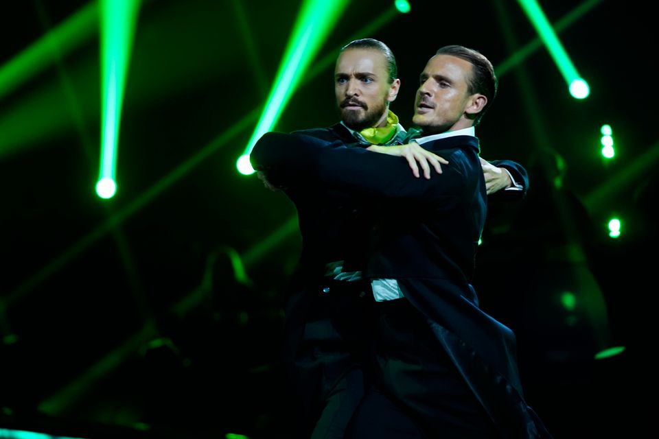 To menn i dress holder hverandre på skuldrene i en tango mens de ser dramatisk fremover.