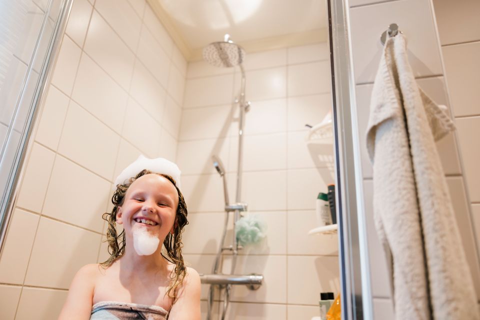 En jente står i dusjen og smiler med et håndkle rundt seg og hvitt skum på hodet og haken.