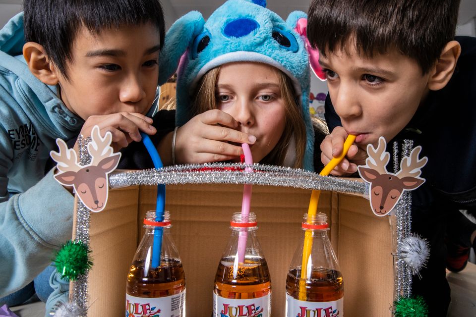 Tre barn lener seg over sugerørene på pappesken og drikker, inni pappesken står tre brusflasker med brun drikke.