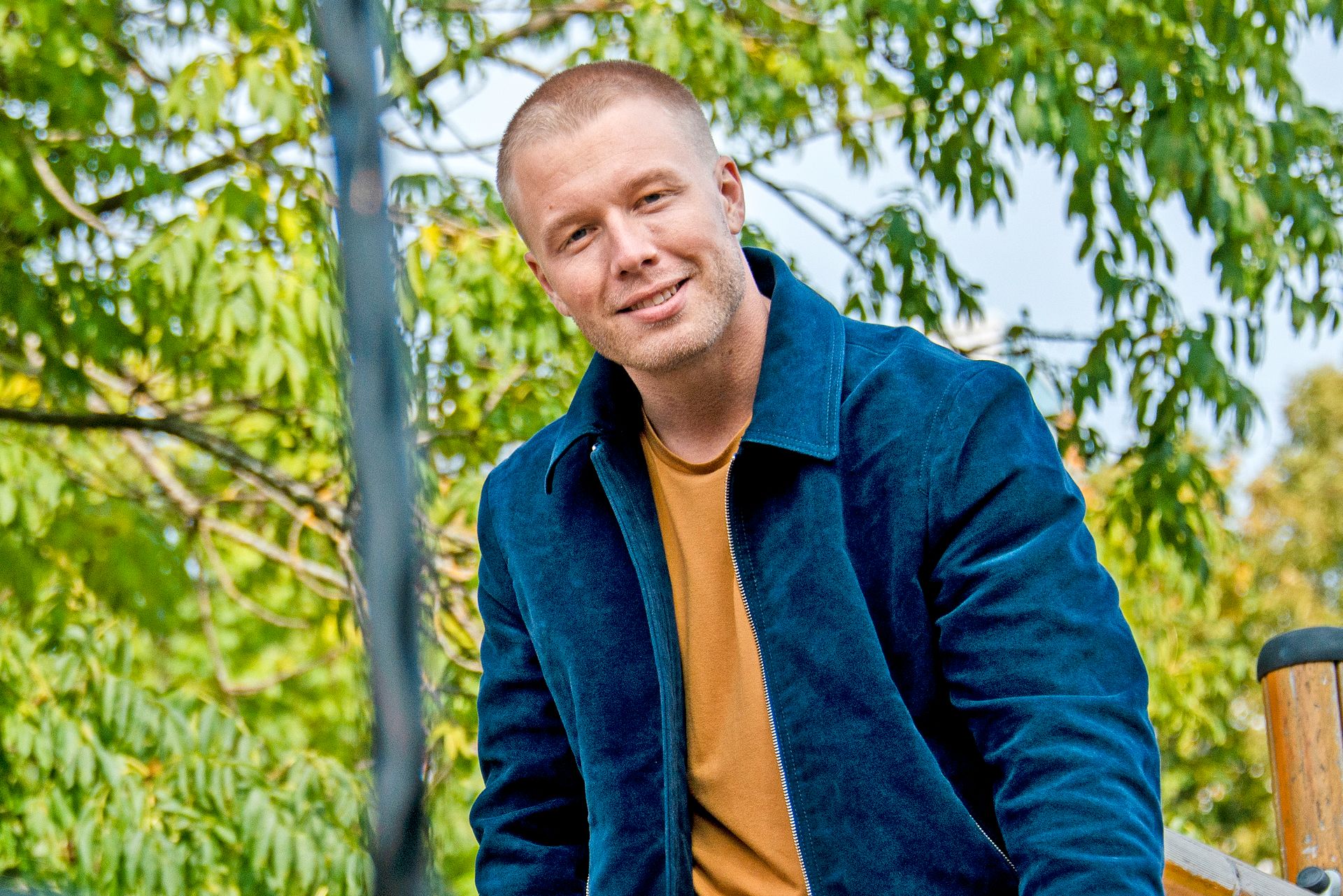 En kortklippet Herman Flesvig har på seg blå jakke og oransje t-skjorte, og smiler mot fotografen, ute i en grønn park.