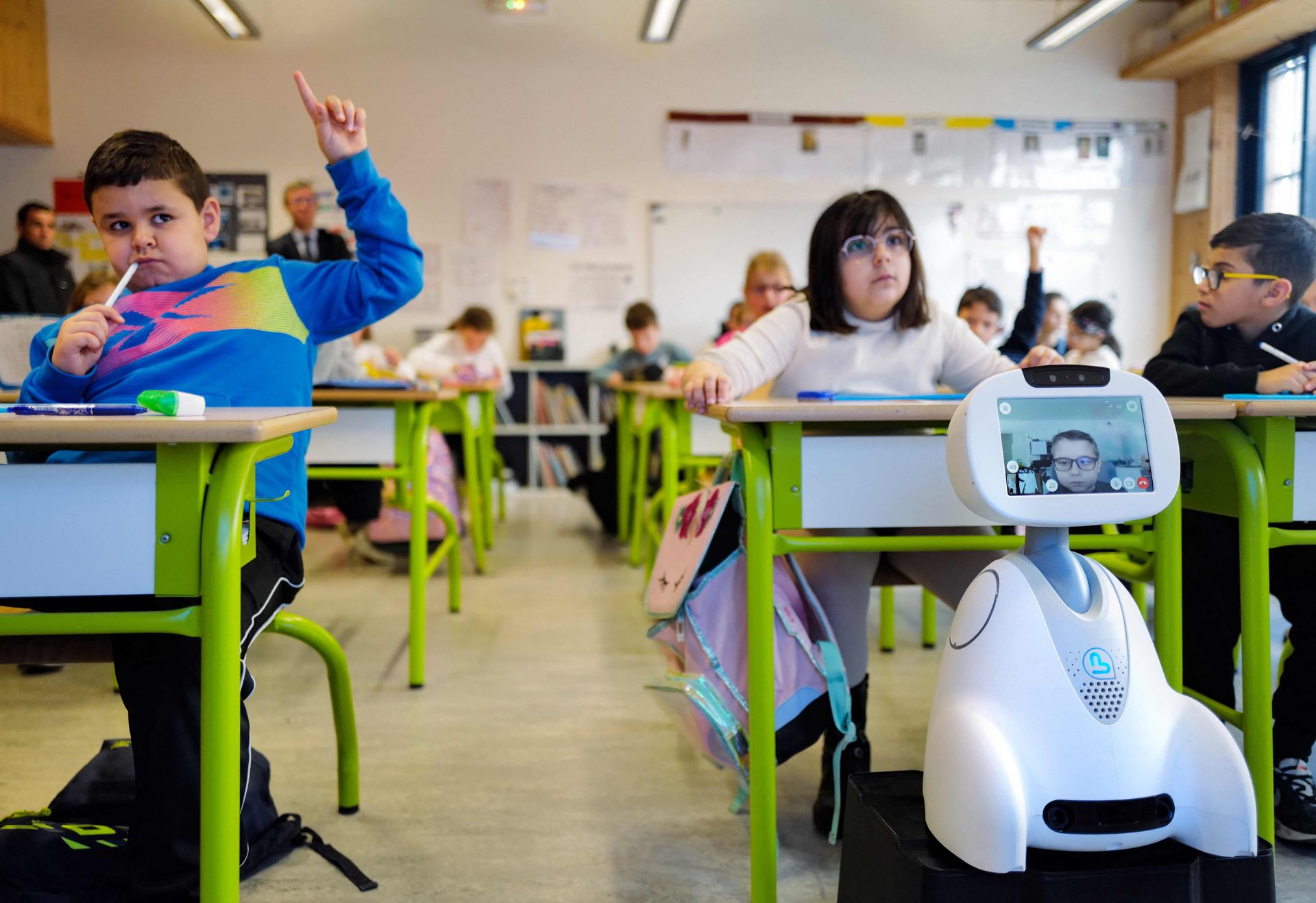 Flere elever sitter bak grønne pulter og rekker hånden i været, og foran den ene eleven står det en liten, hvit robot på et bord, med en skjerm som viser ansiktet til en elev.
