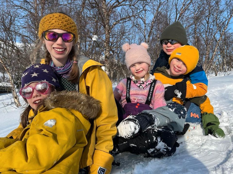 Fem barn mellom seks og tolv år, alle med gule og ellers fargerike vinterklær på, sitter sammen ute i snøen og smiler.