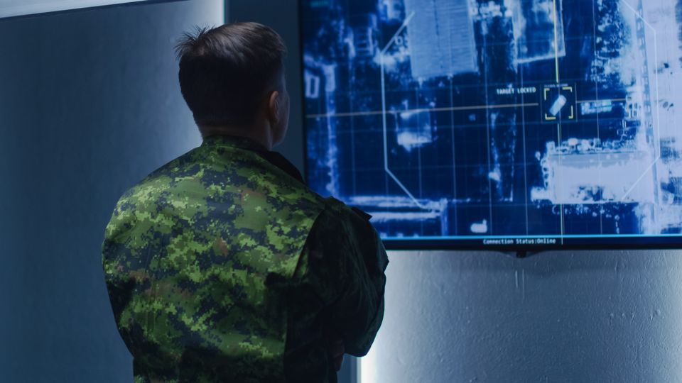 En illustrasjon viser en mann i kamuflasjeklær som står med ryggen til og ser på en skjerm med et blått satellitt-kartbilde.