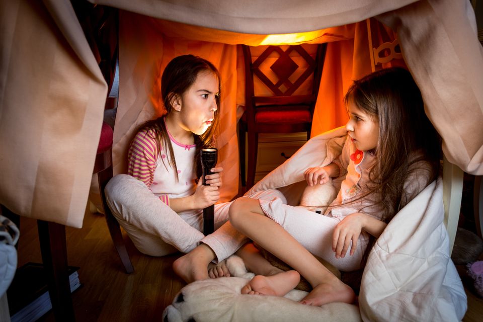 Tre jenter sitter under et bord med tepper og laken over, så det blir som et telt, og lyser opp ansiktet sitt med lommelykt og ser litt redde ut.