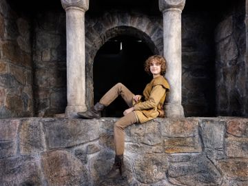 En jente med middelalderklær og kort, krøllete hår sitter på en steinmur og smiler.