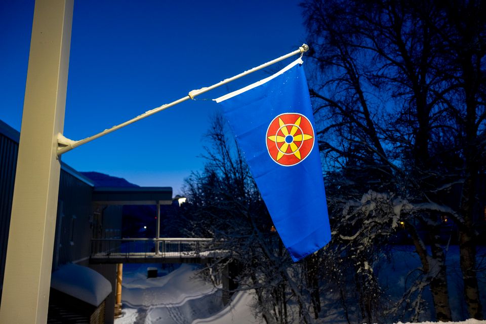 Det kvenske flagget henger ved en ytterdør med nattehimmel og trær i bakgrunnen. Flagget er blått med en runding med en seksbladet rose.