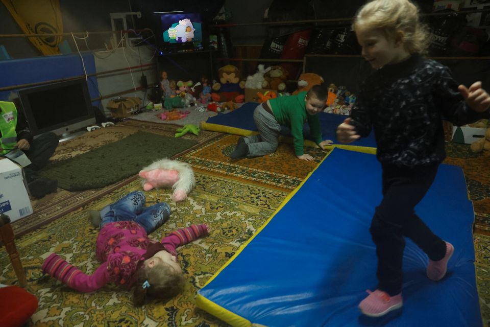 Flere små barn leker inne på et rom - ei jente løper på en blå madrass mens ei annen ligger på gulvet ved siden av og en gutt med grønn genser krabber.