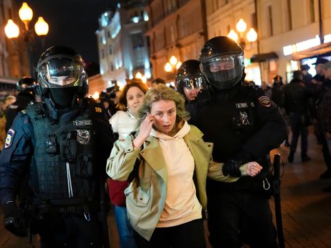 En kvinne med grønn ytterjakke blir ført bortover en gate av to store politimenn i svarte vernedrakter og hjelmer med visir. 