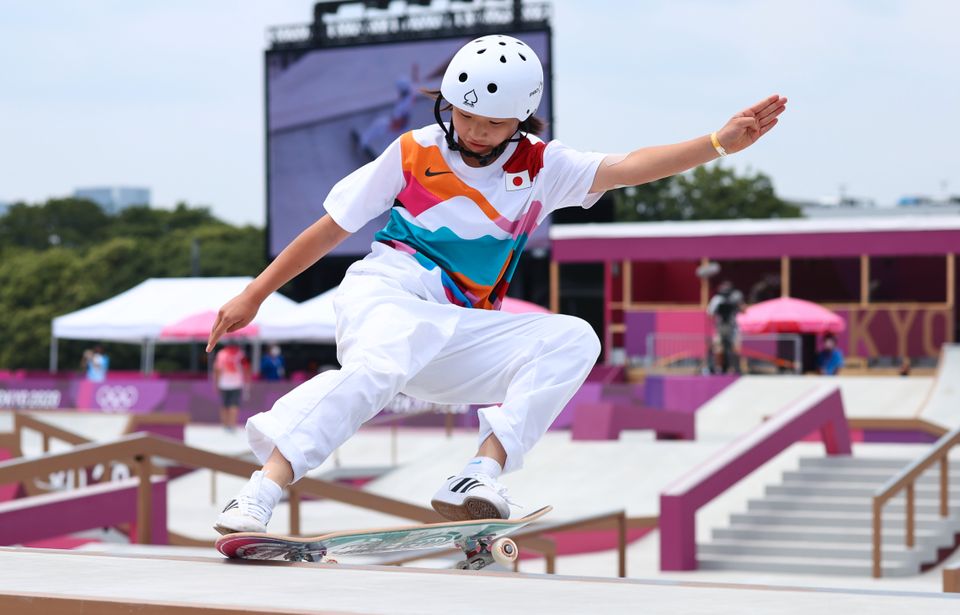 En ung jente med hvite klær med japansk flagg på, hjelm, sneakers og skateboard er fryst i bevegelse på kanten av en OL-skaterampe.
