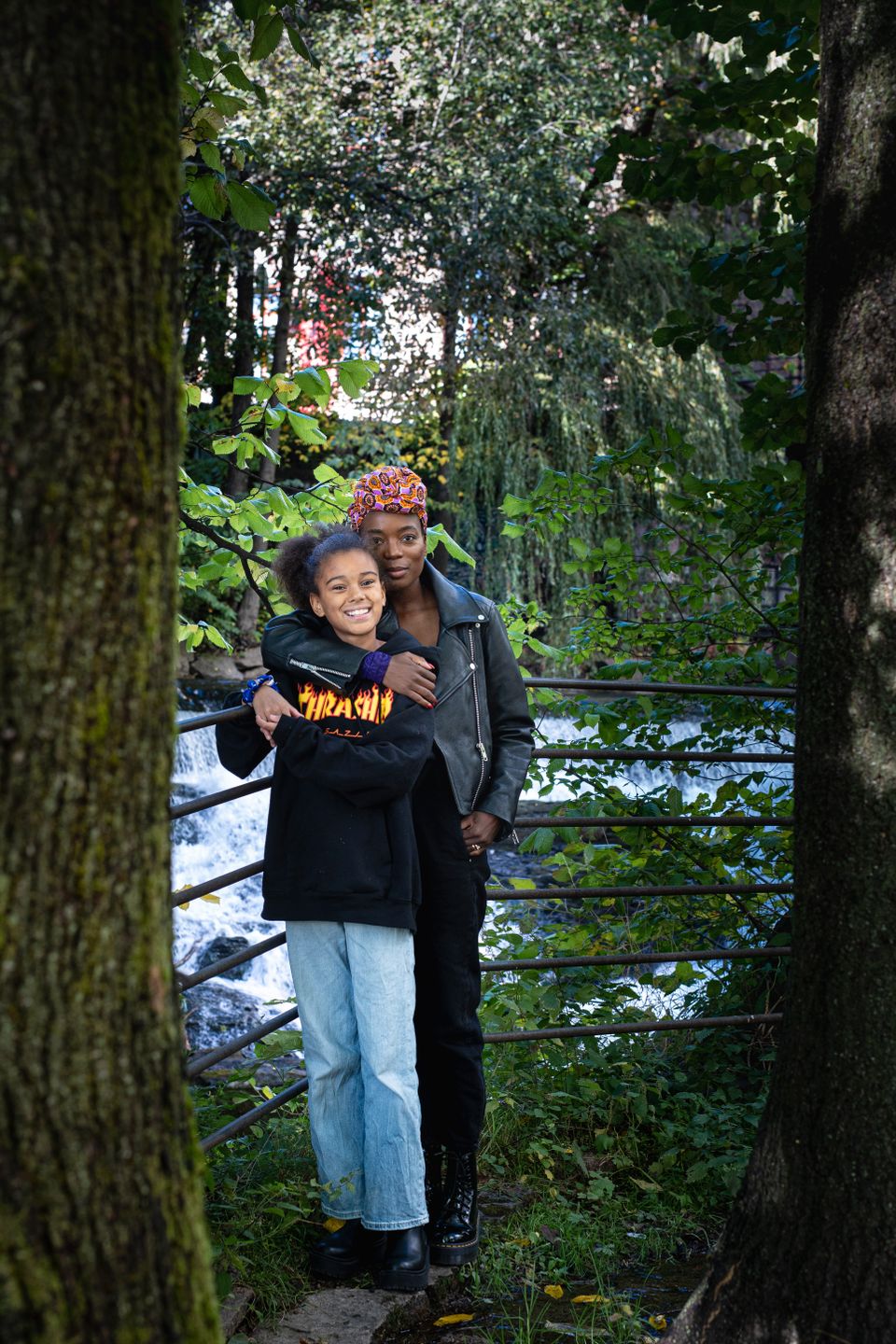 En voksen dame står og holder rundt en ung jente mellom to trær med en elv i bakgrunnen.